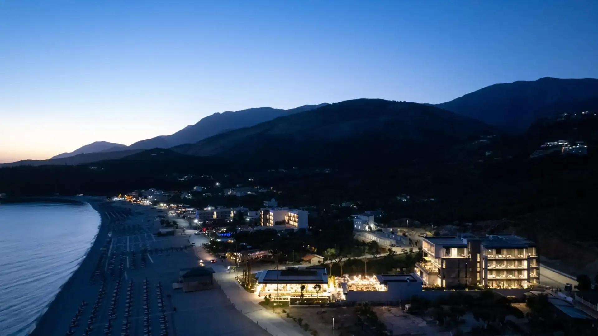 Livadhi bay from the air at dusk with the lit buildings along the shore and the mountains standing behind