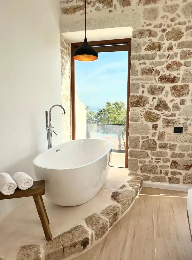 A white freestanding bath below a black pendant lamp, standing on a pale plaster dais edged with rough stone, beside a full-height glazed door onto a balcony above the sea, with white plaster to the left and rubble limestone to the right.