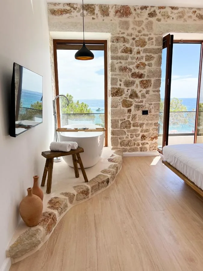 A freestanding bath on a stone-kerbed plaster dais, two terracotta amphorae standing at the near end of the same dais under a wall-mounted television that reflects the sea, and two openings onto the water.