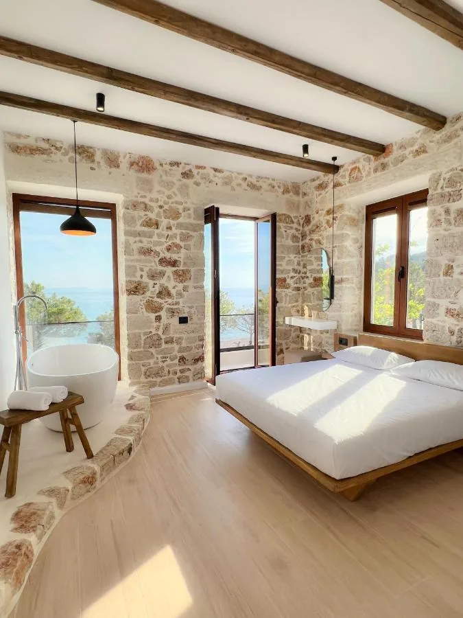 A stone-walled room under squared timber ceiling beams, with a freestanding oval bath on a stone-kerbed dais to the left, a low timber platform bed to the right, and three openings — a window and a balcony door onto the sea, and a two-leaf window onto the trees behind.