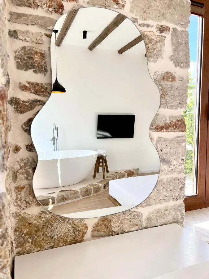 A large frameless mirror with a wavy scalloped edge hung flat against a rubble limestone wall above a white plaster ledge, reflecting the ceiling beams, the pendant lamp, a wall-mounted television, the bath and the corner of the bed.