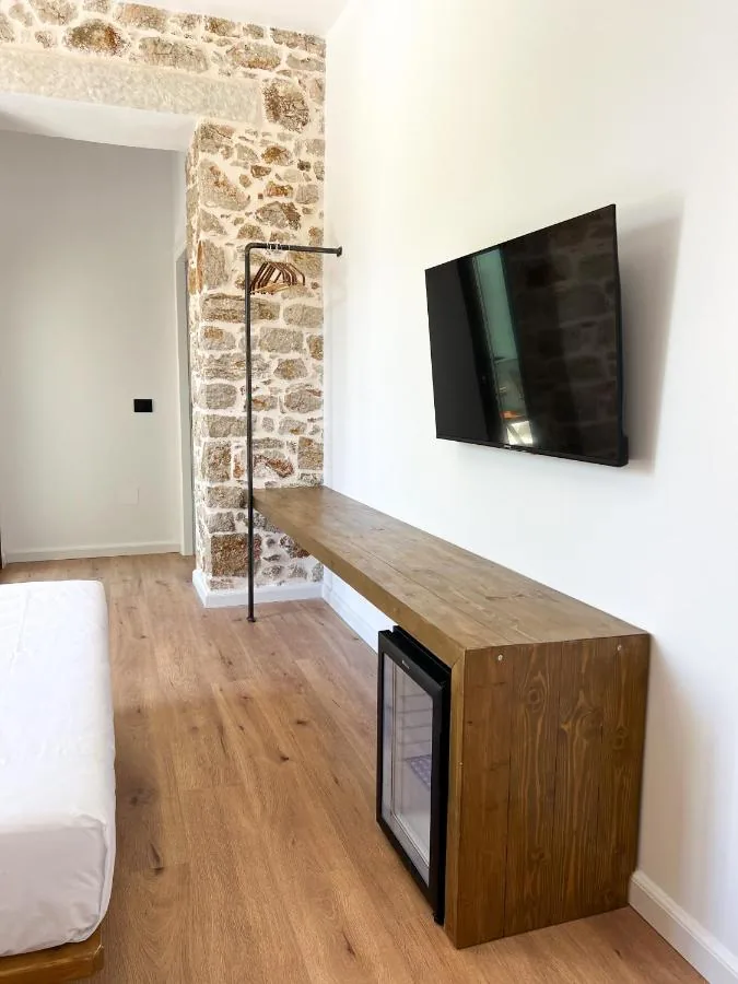 A room with white plastered walls and one exposed limestone pier, a wide oak plank floor, a long timber shelf running under a wall-mounted television, a glass-fronted fridge beneath it and a black hanging rail.