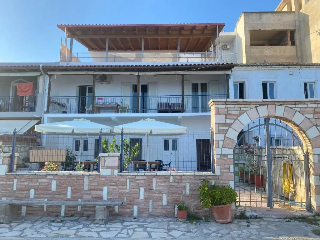 The full white front of the building in summer, two floors of balconies with slate-grey rails, parasols on the terrace and the timber pergola on the roof
