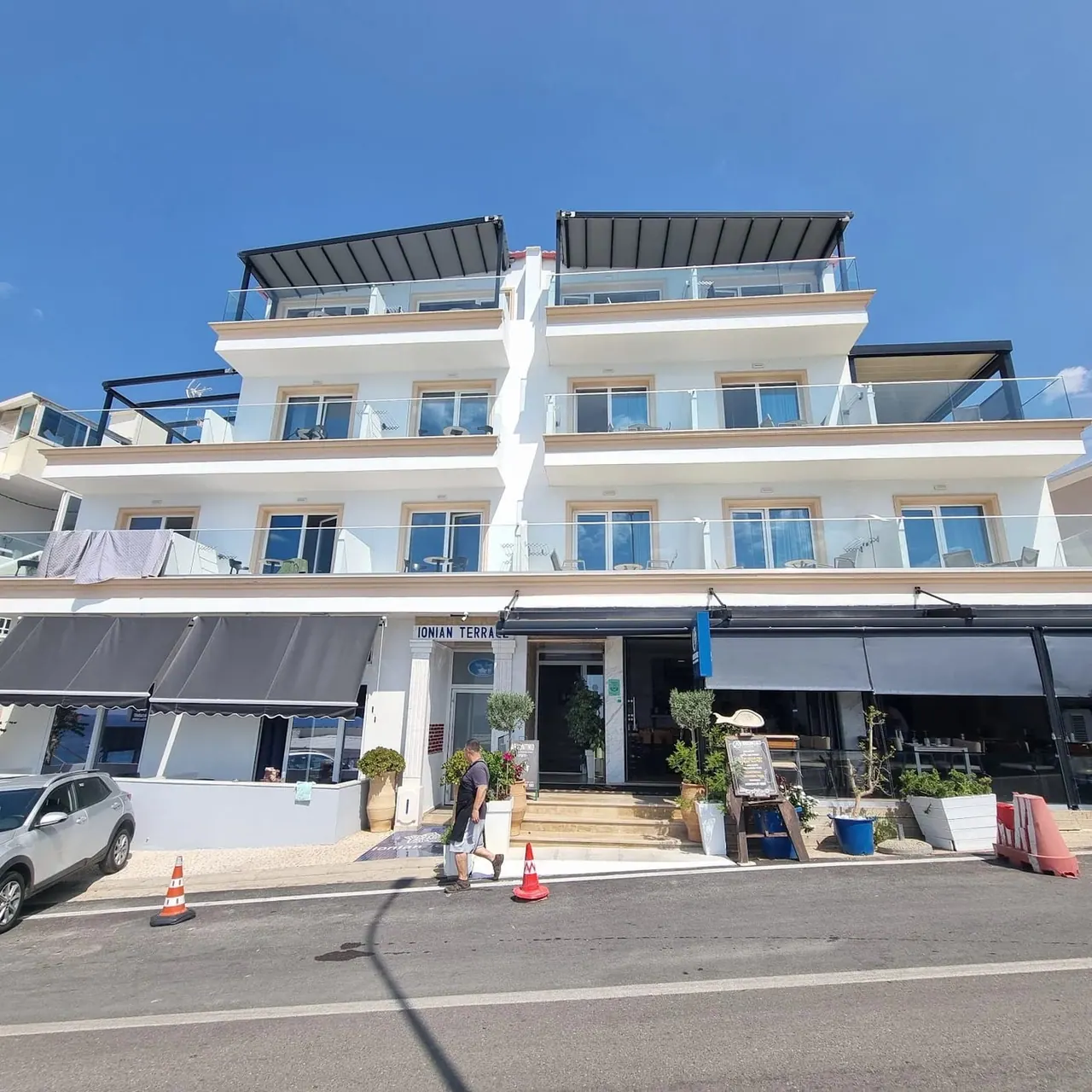 The building from the seafront road, with dark awnings over the ground floor, potted olives and blue planters along the pavement