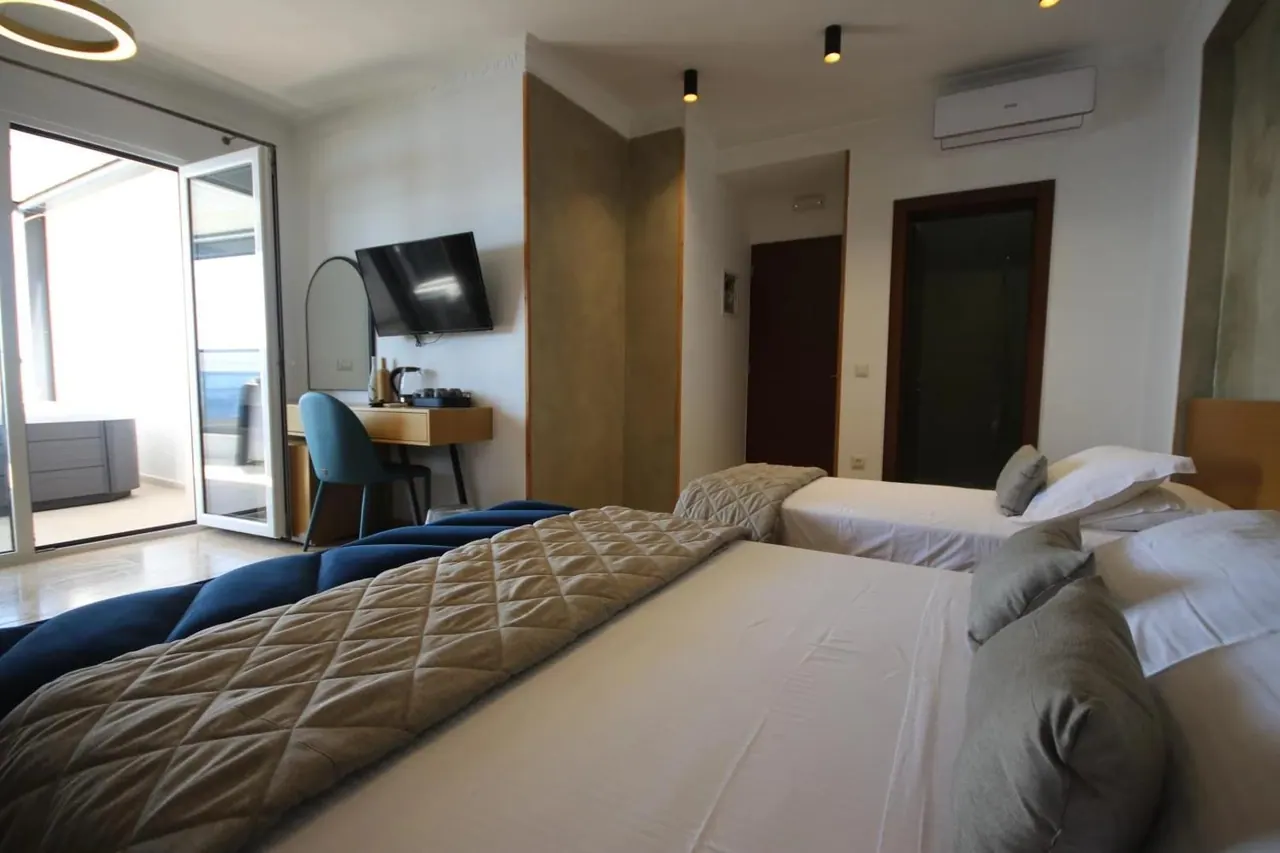 A bedroom with its glazed door swung open onto a terrace, a desk and mirror along the wall and a blue bench at the foot of the bed
