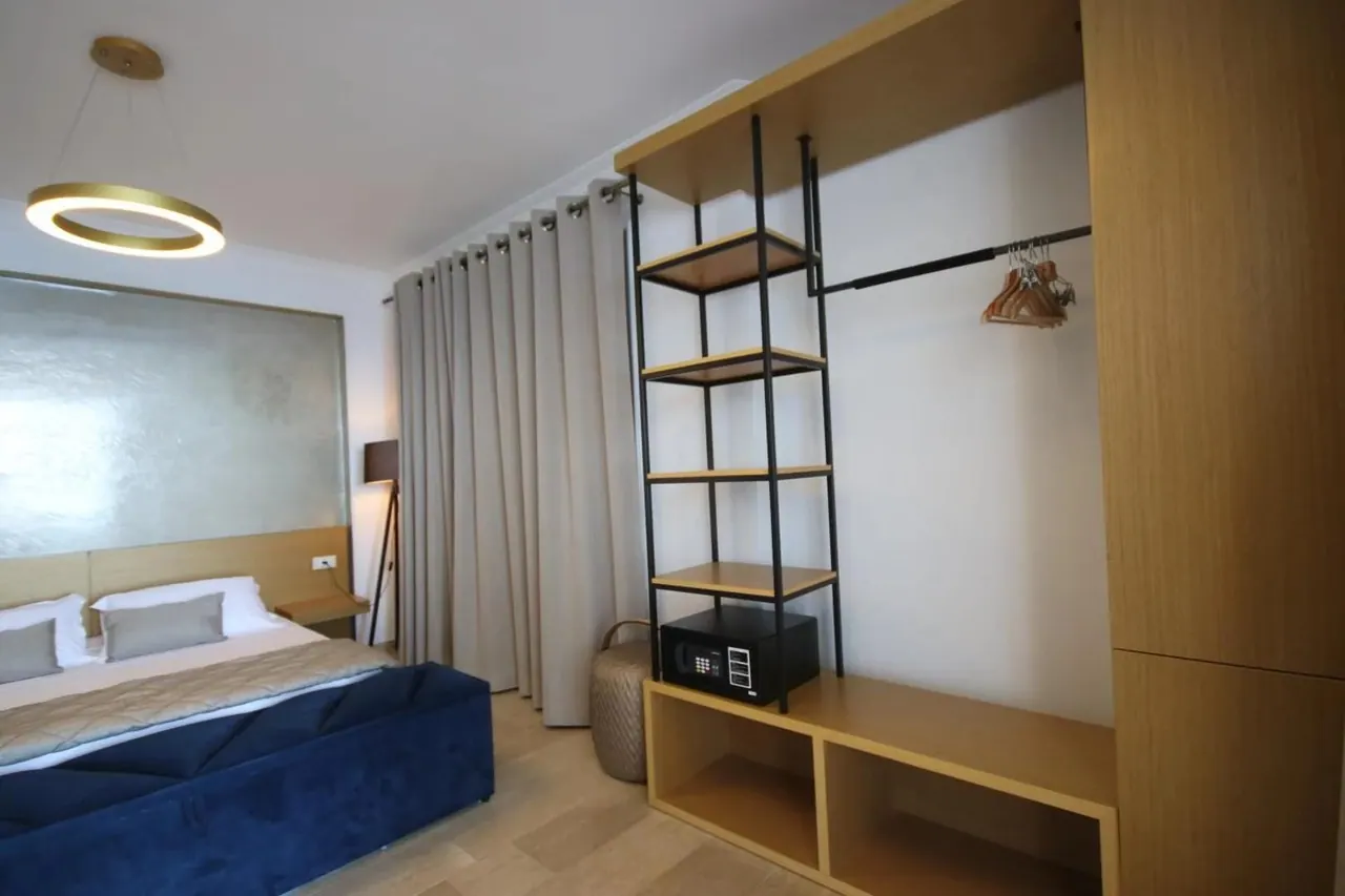 A bedroom with a timber wardrobe, black steel shelving holding a safe, and a blue upholstered bench at the foot of the bed