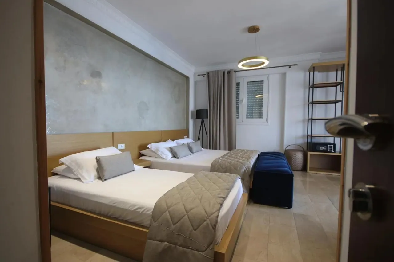 A room seen from its open doorway: two beds under quilted throws, a blue velvet bench, a steel shelving bay and a shuttered window