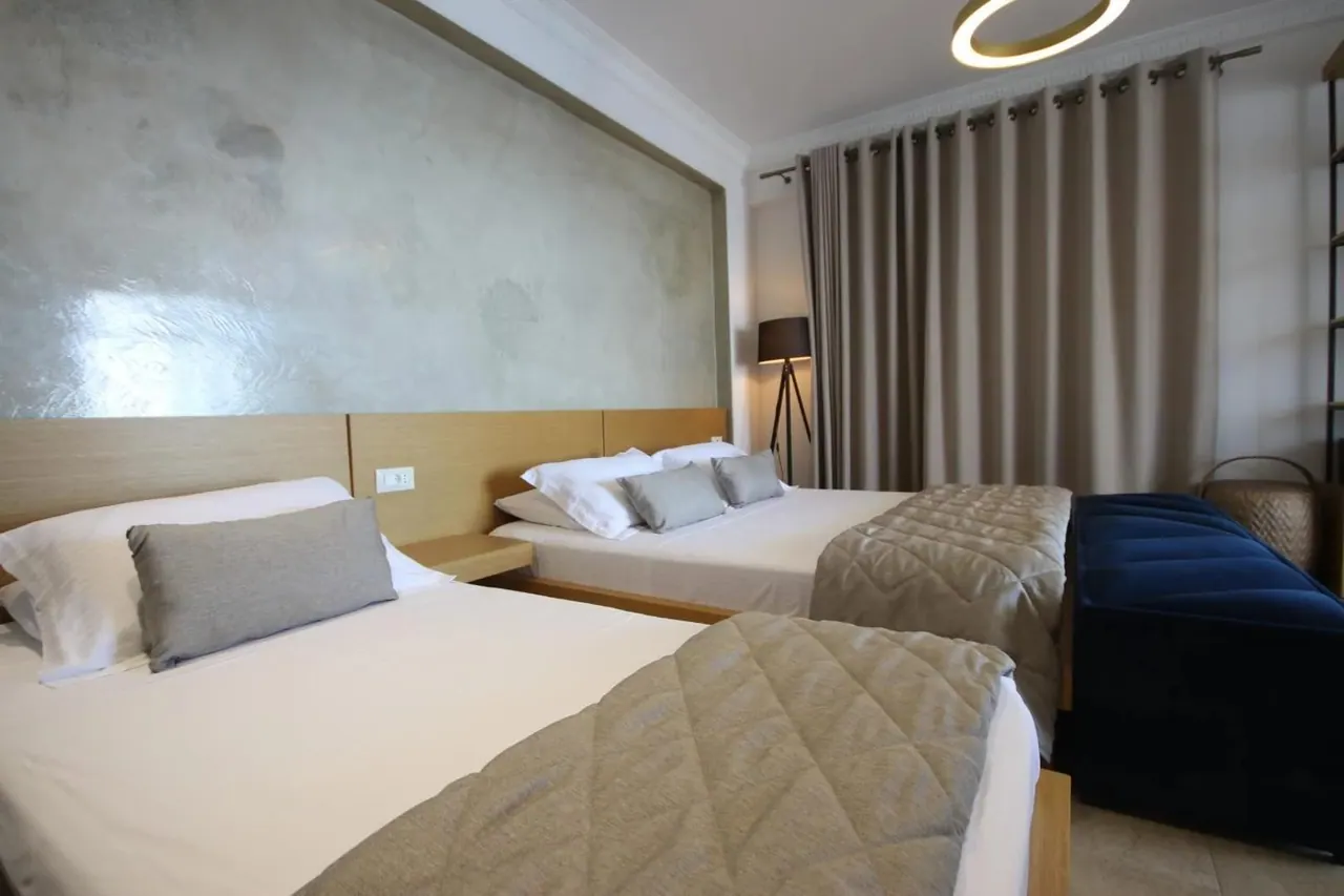 Two beds side by side under quilted throws, with timber headboards under a panel of polished plaster and a standing lamp