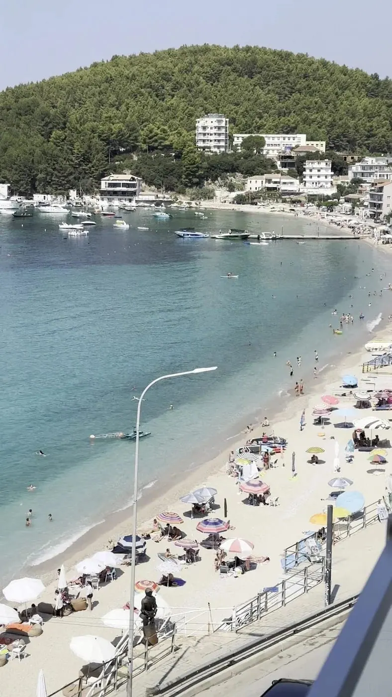 The bay from an upper floor: the sandy town beach with its umbrellas, moored boats and the pine headland closing the far side