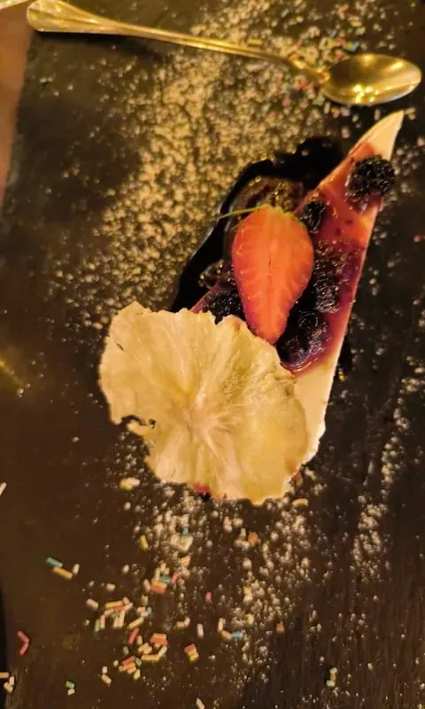 A wedge of pale set dessert under dark berry compote and a whole blackberry, with a halved strawberry and a large dried pineapple slice, on dark slate strewn with sugar and coloured sprinkles beside a gold spoon