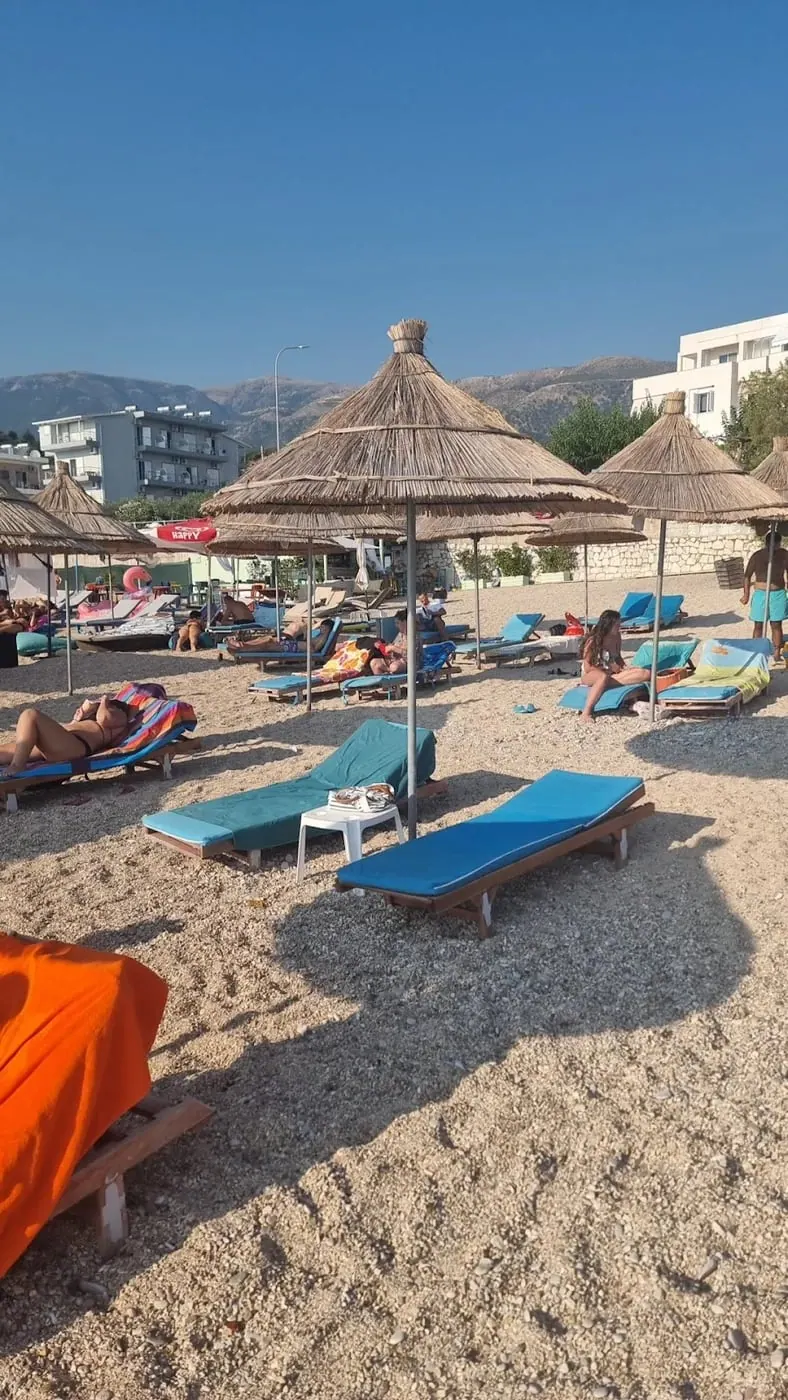 Blue sunbeds and thatched umbrellas on grey pebbles, with the coast road and the hills behind