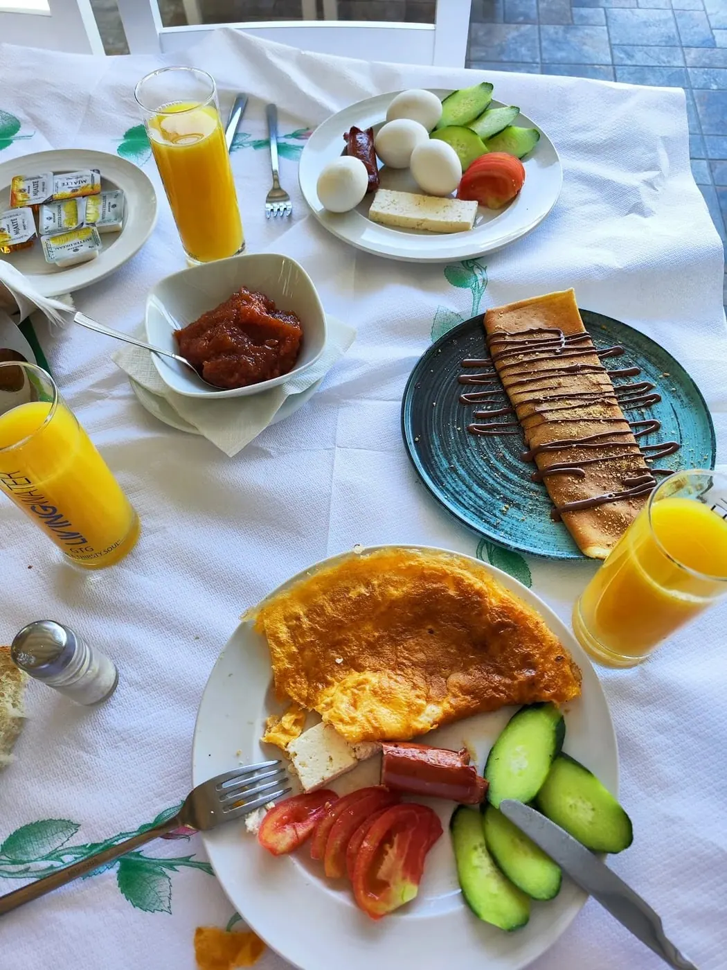 A breakfast table with omelette, a chocolate crêpe, boiled eggs, cheese, tomato, cucumber and orange juice