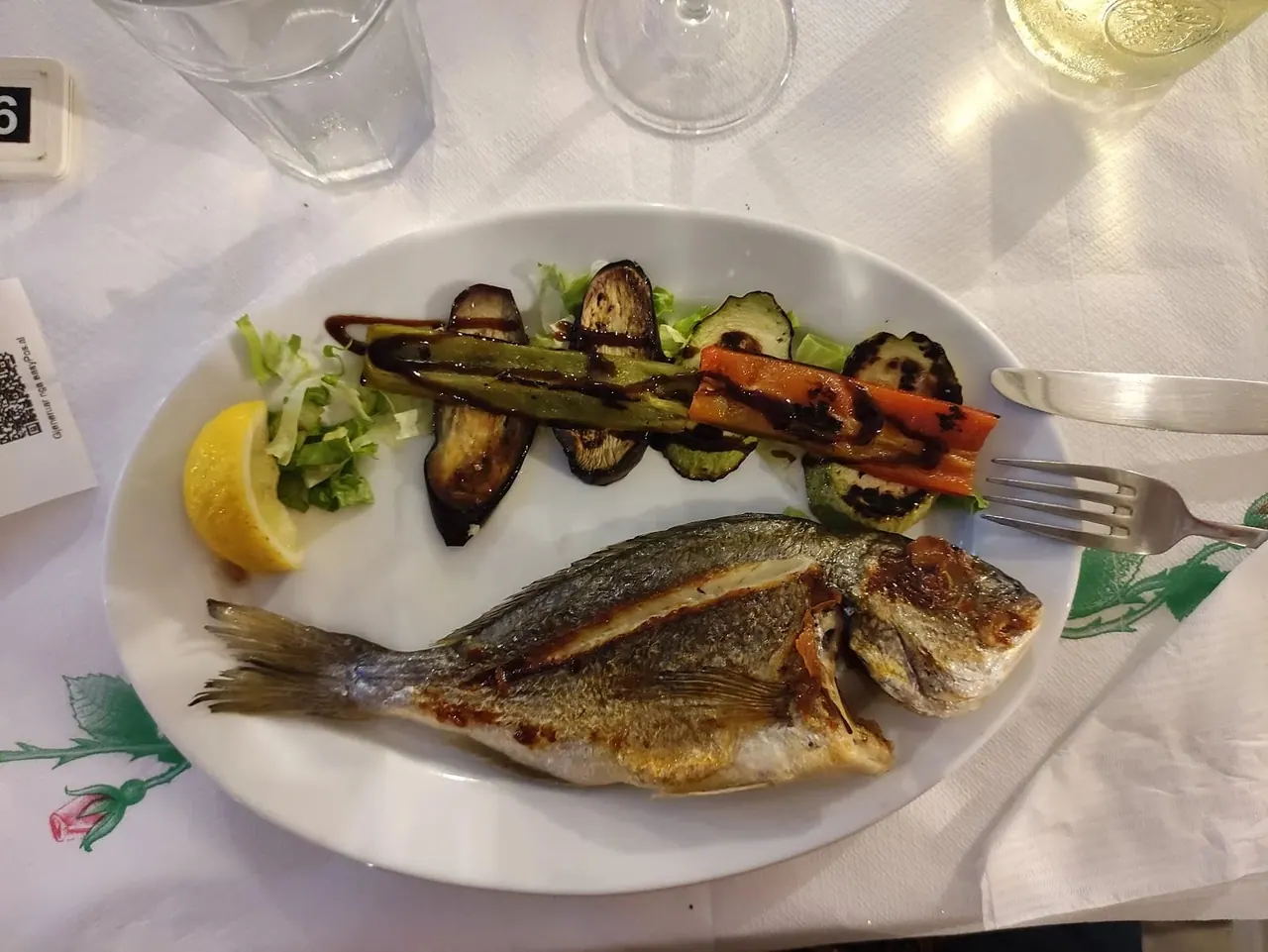A whole grilled sea bream on a white plate with charred aubergine, pepper and lemon