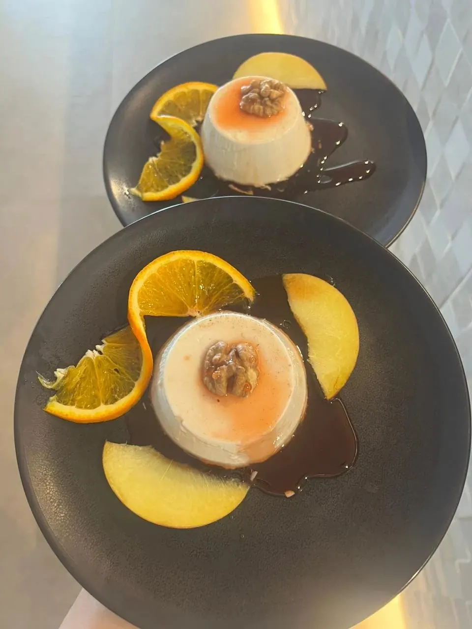 Two panna cottas on dark plates, each under a walnut half, with orange slices and a dark syrup