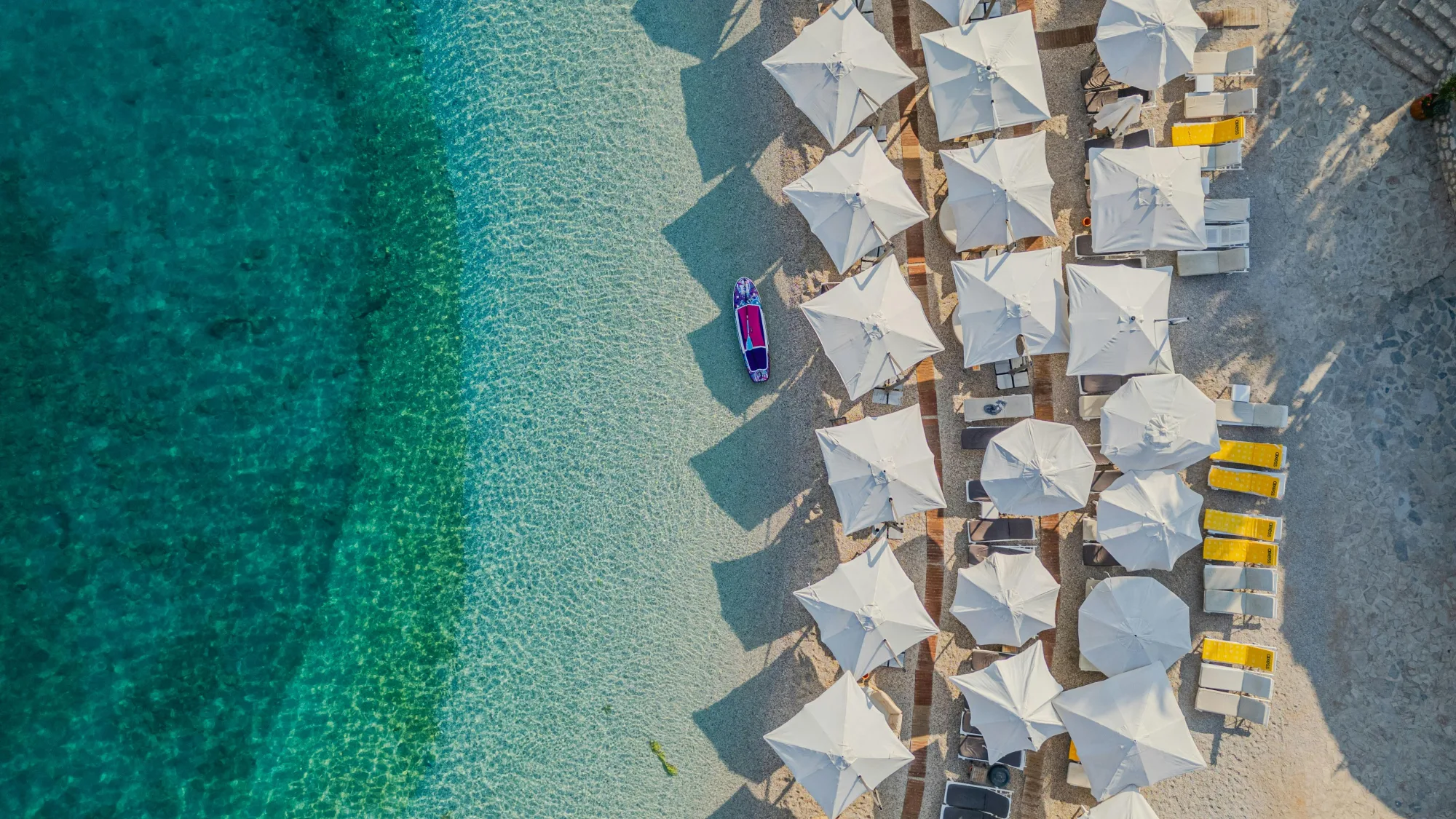 Aerial view of Dhermi Beach with rows of white sunbed umbrellas and a SUP board on turquoise Ionian water