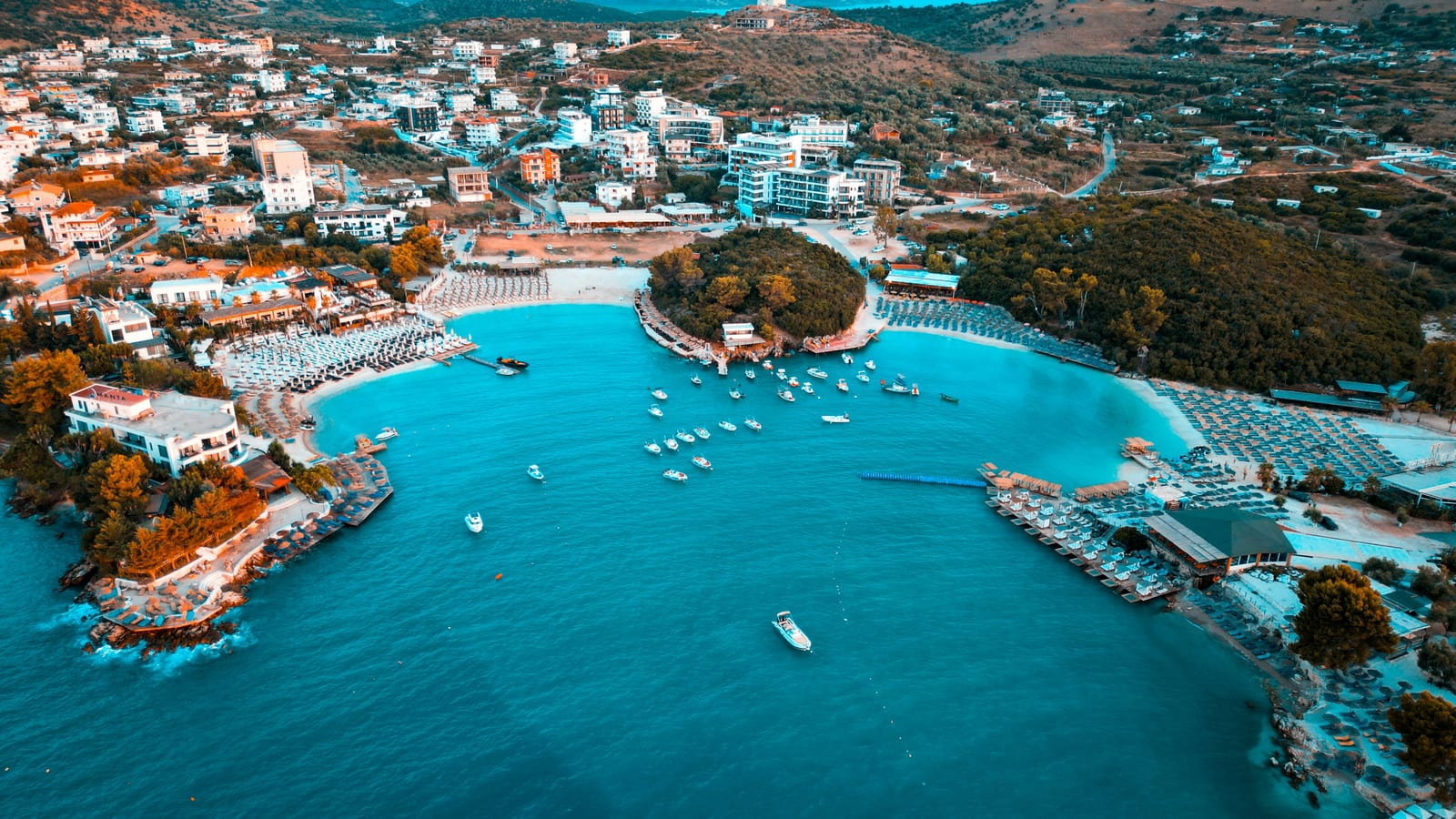 Aerial of Ksamil town and beach clubs at golden hour — the resort bay where most hotels and apartments cluster