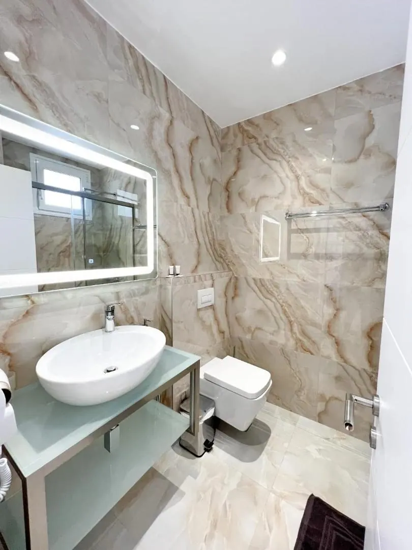 A bathroom lined in caramel onyx tile, with a vessel basin on a glass shelf and a lit mirror