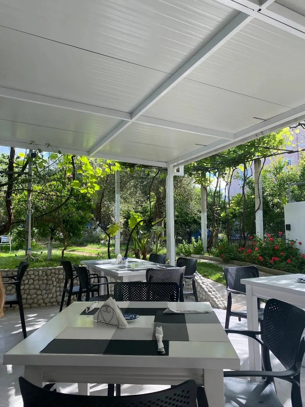White tables set for breakfast under the garden canopy, grape vines and olive trees beyond a pebble-faced wall