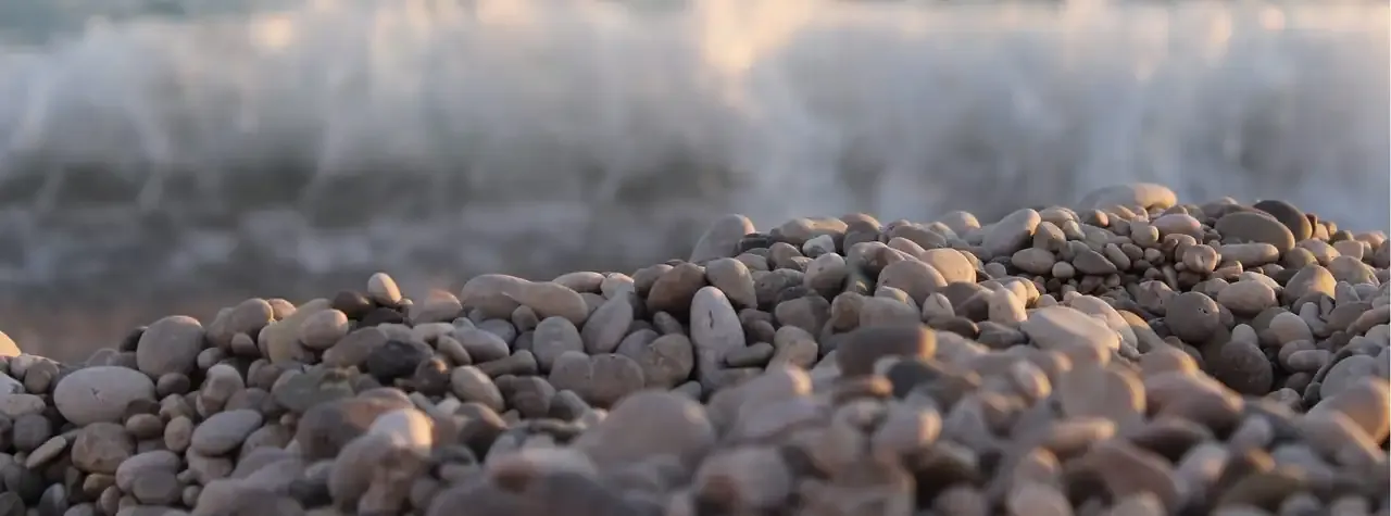 The pebbles of Livadhi beach in close-up, warm tan and grey, the surf breaking behind them