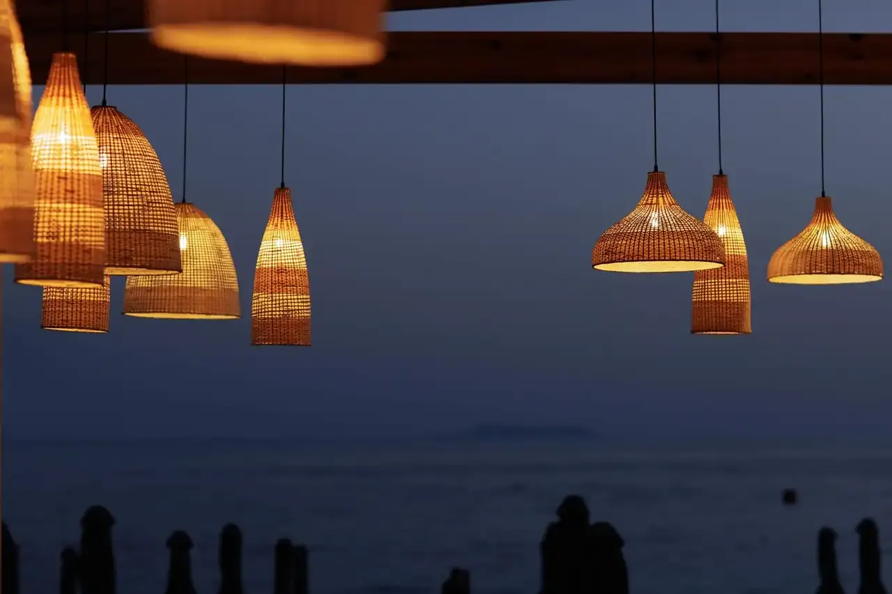 Rattan pendants lit above the beach bar at dusk, the Ionian dark behind them