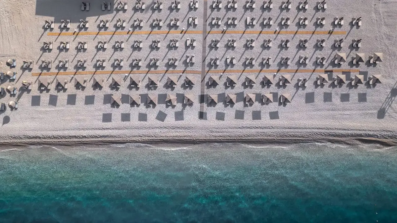 The hotel's beach from directly overhead, straw umbrellas in ruled rows on pale shingle with the shoreline running across the frame
