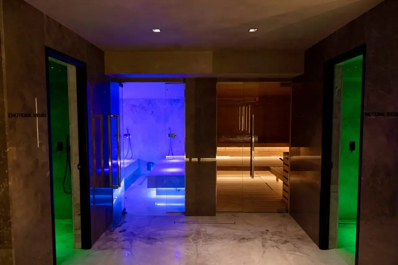 The wet area of the spa at Prado: two green-lit shower cabins, a blue-lit steam room between them and the timber sauna through glass