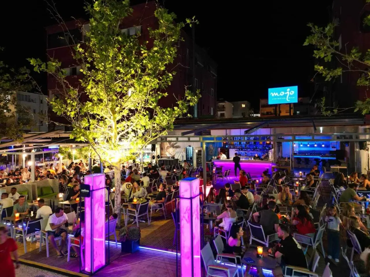 Busy evening terrace at Mojo Cocktails Bar with the illuminated Mojo sign and central bar in Spile