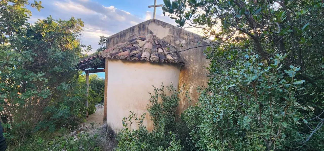 Small whitewashed Orthodox chapel of Saint Nicholas with a stone-tiled roof and cross, surrounded by greenery near Dhërmi