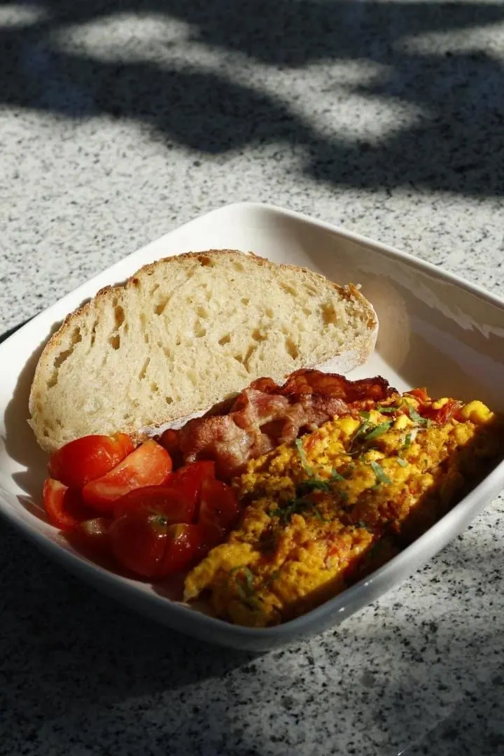 Scrambled eggs with pancetta, halved cherry tomatoes and a thick slice of bread on a stone table