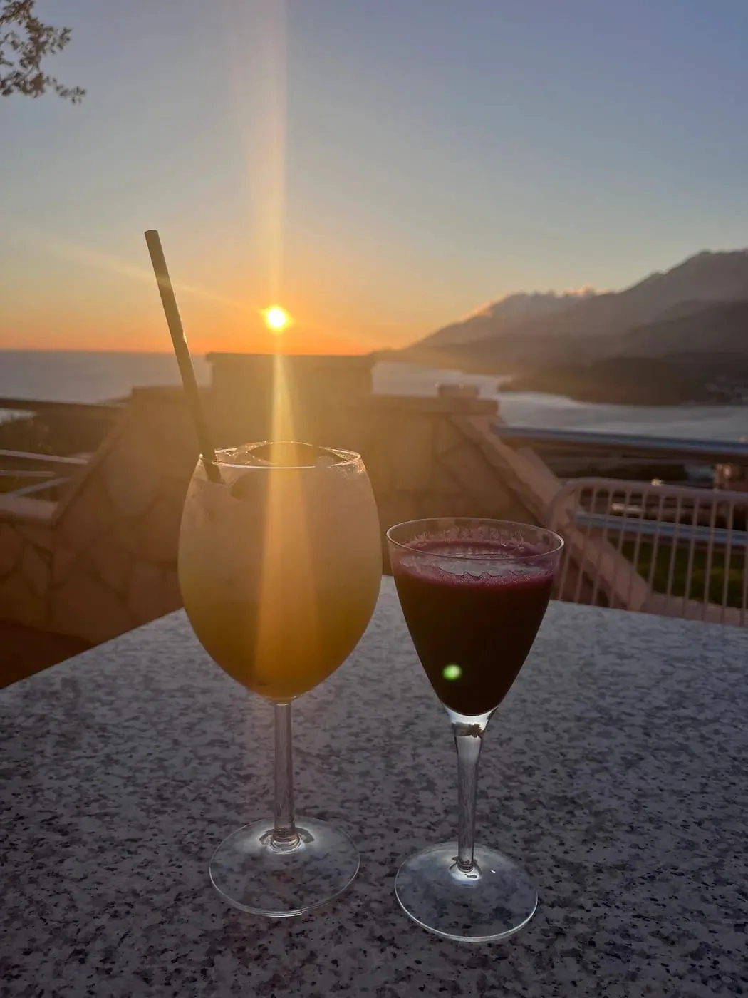 Two long drinks on a stone table with the sun setting over the sea behind them