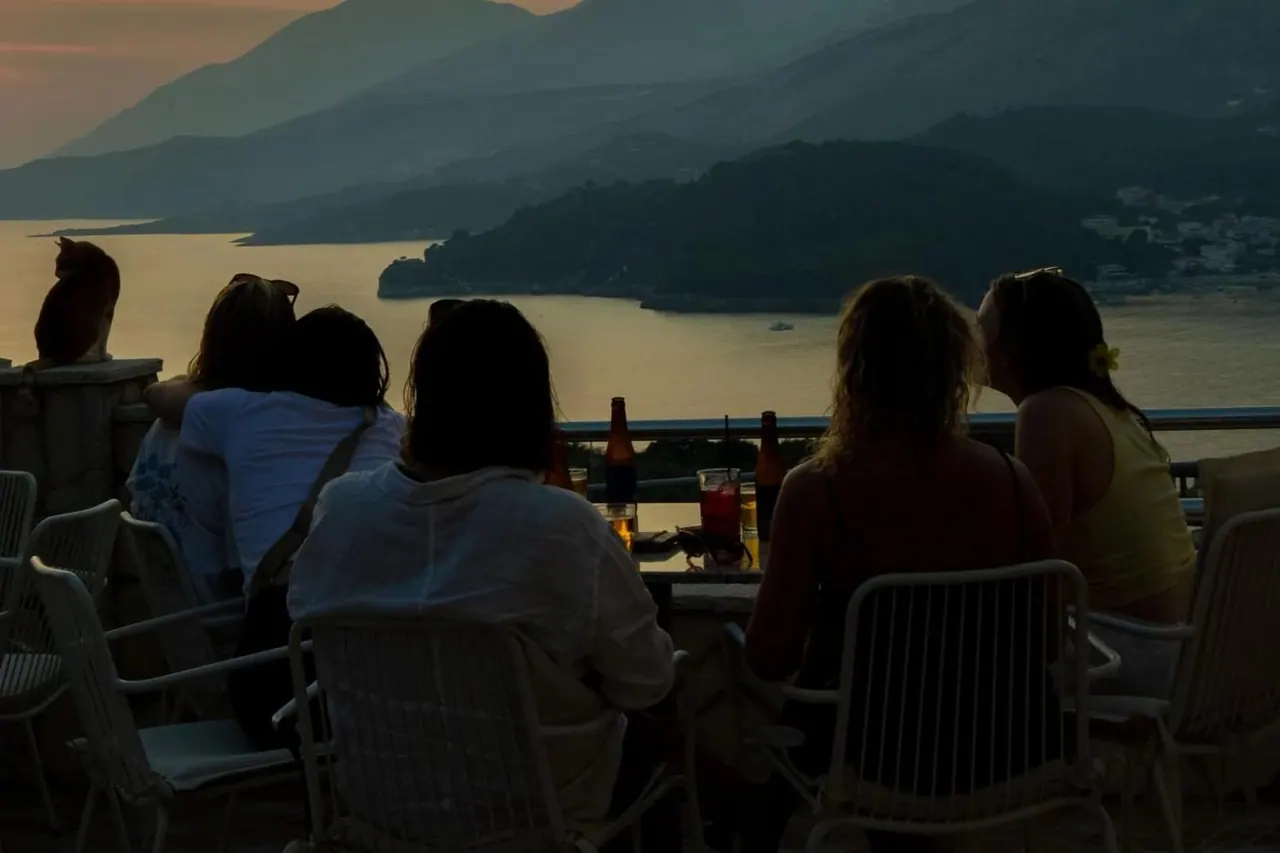 Guests seated at the terrace rail watching the sun set over Himarë bay, beer and a spritz on the table, a cat on the wall