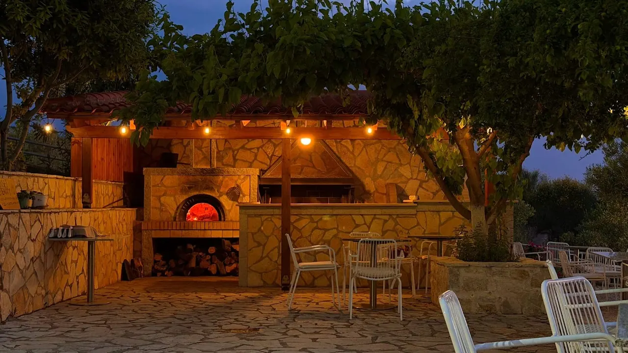The stone wood-fired oven glowing under its tiled timber pergola at dusk, festoon bulbs lit above