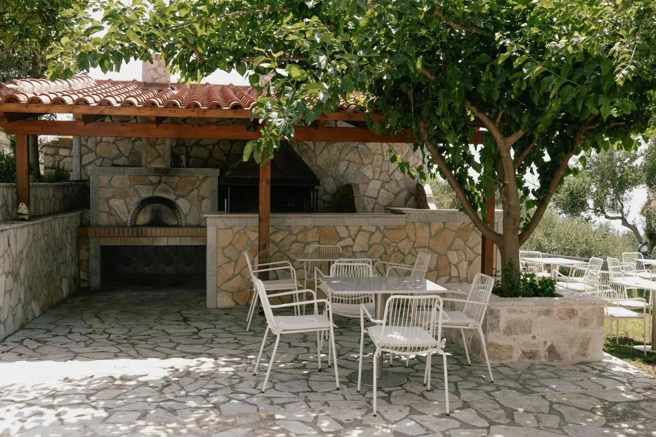 The oven and its stone counter in daylight with white chairs and tables under a broad-leaved tree