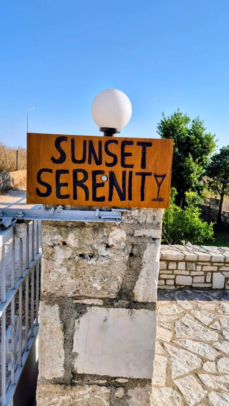 A hand-painted timber sign reading SUNSET SERENITY on a stone gatepost, the Y drawn as a martini glass
