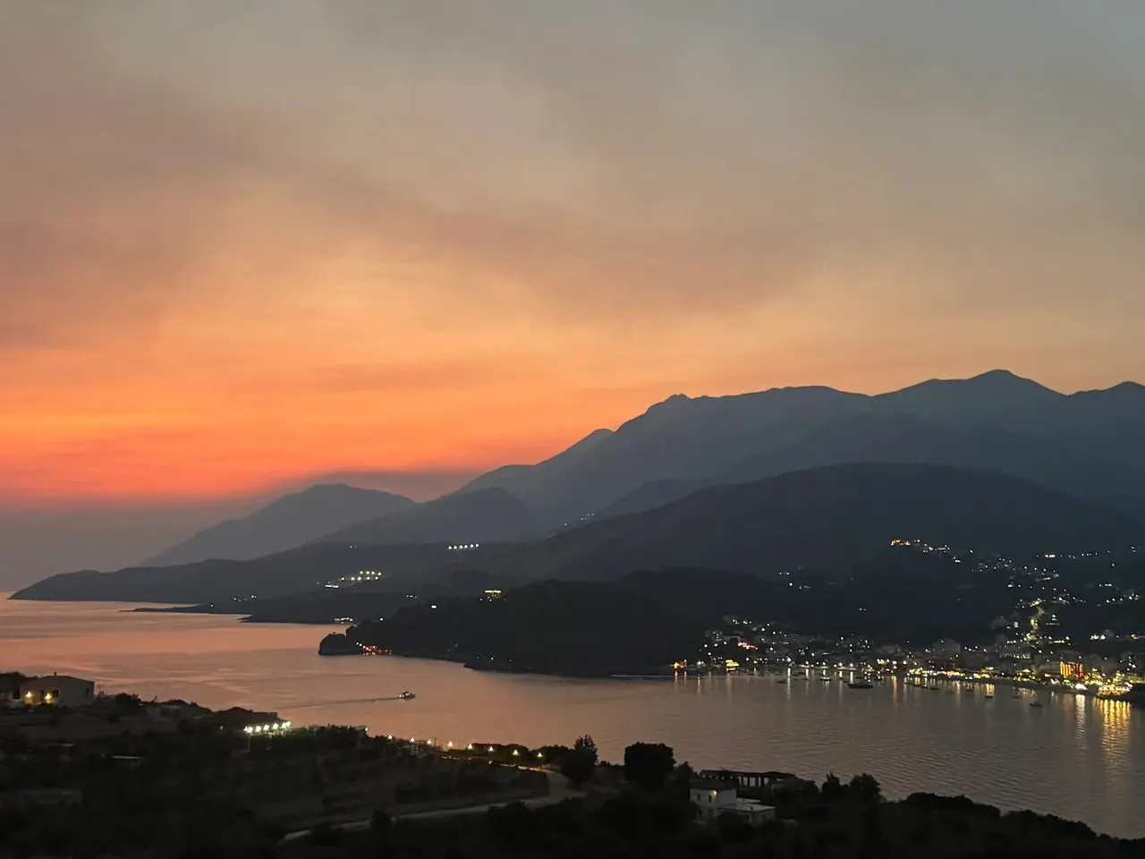 Himarë bay after sunset, orange banding the sky over dark headlands and the town lit along the shore