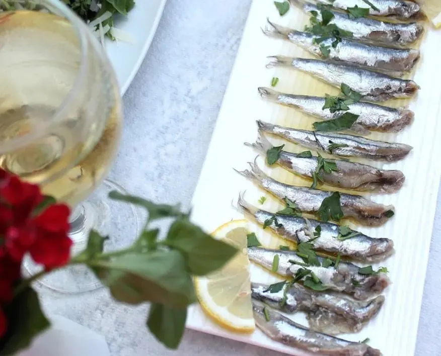 A long white plate of anchovies dressed with lemon, olive oil and chopped parsley, beside a glass of white wine