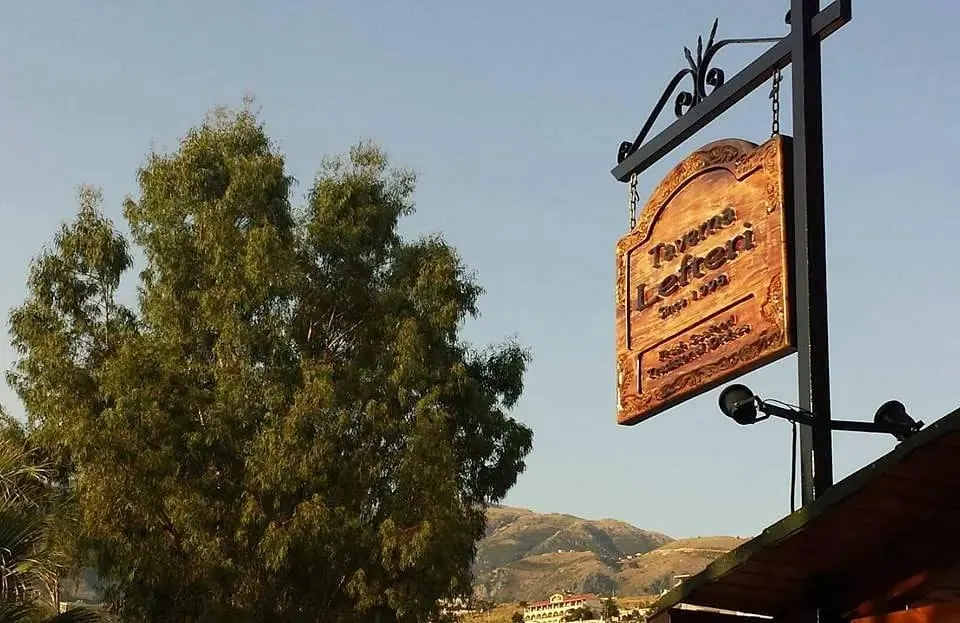 The carved oak sign of Taverna Lefteri hanging from a wrought-iron bracket, with the Himarë hills behind