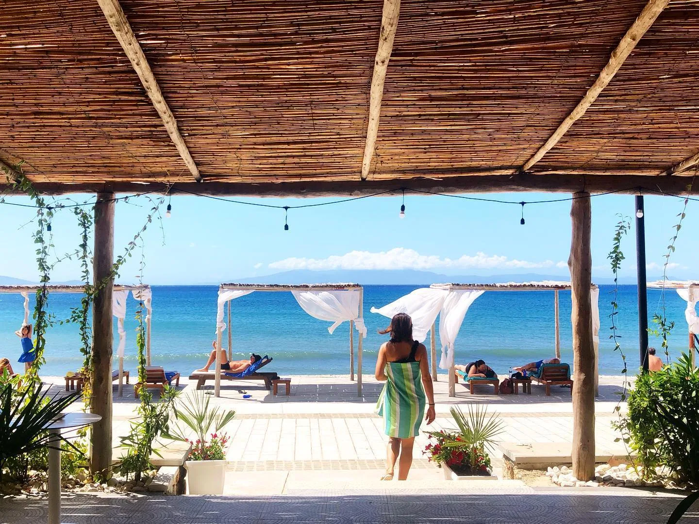 White-curtained daybeds on a timber deck at the water's edge under a reed roof