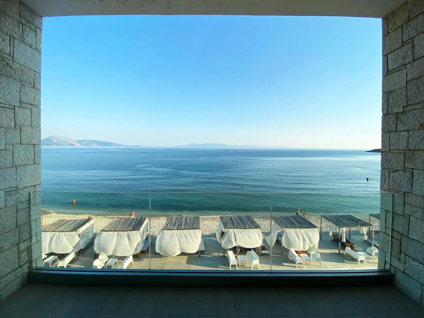 A wide stone-framed opening in the hotel wall looking straight out over a row of white sunbeds to the sea