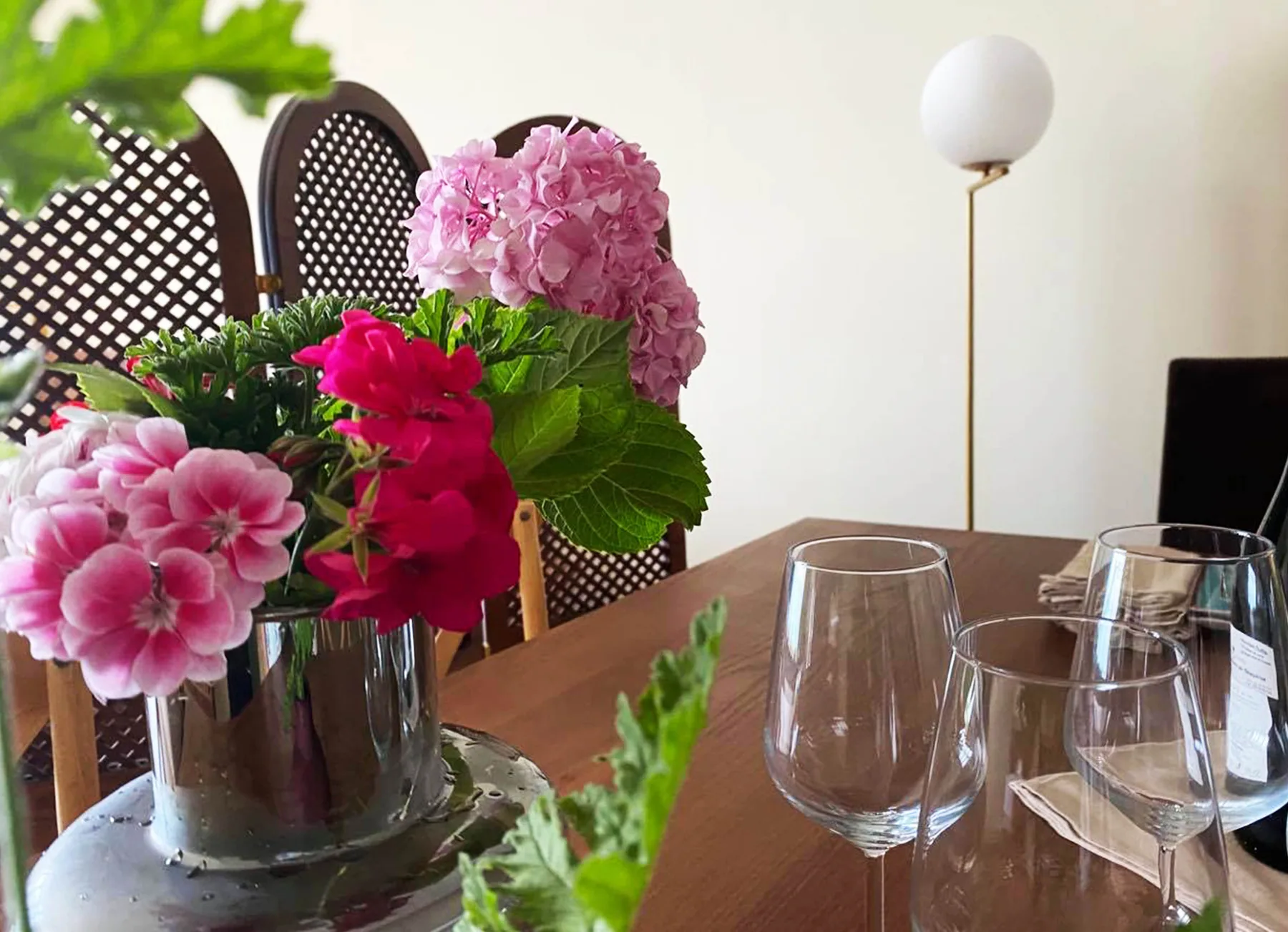 Wine glasses and a jug of pink geraniums on a laid table beside cane-backed chairs