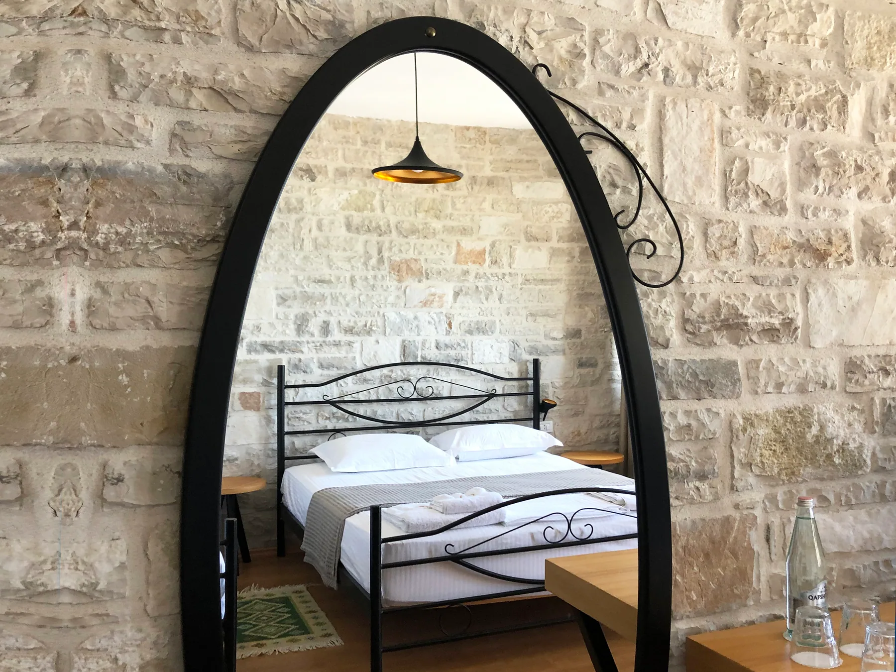 An oval black-framed mirror on a rough limestone wall reflecting a made bed