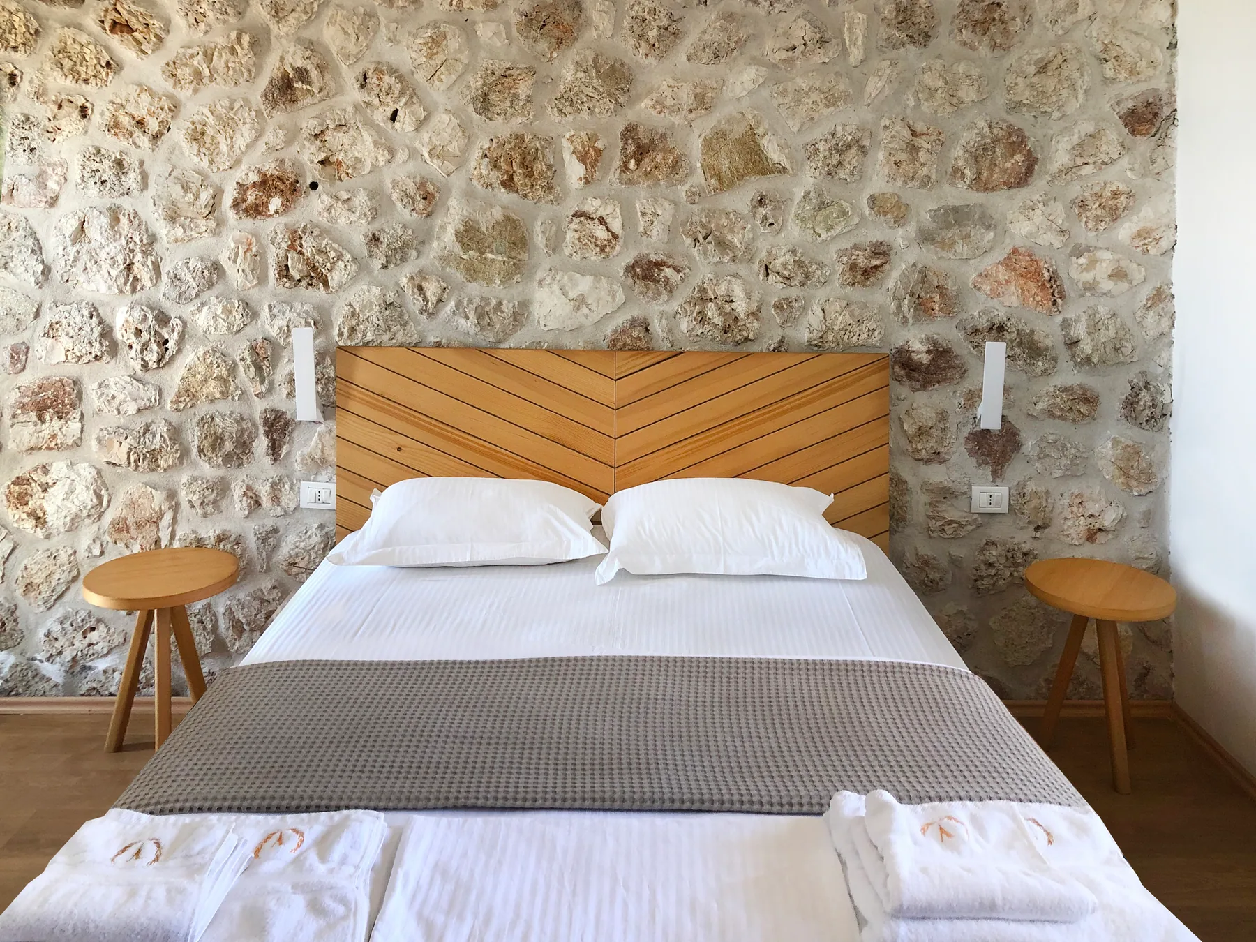 A double bed with a chevron timber headboard against a limestone wall, a round stool either side