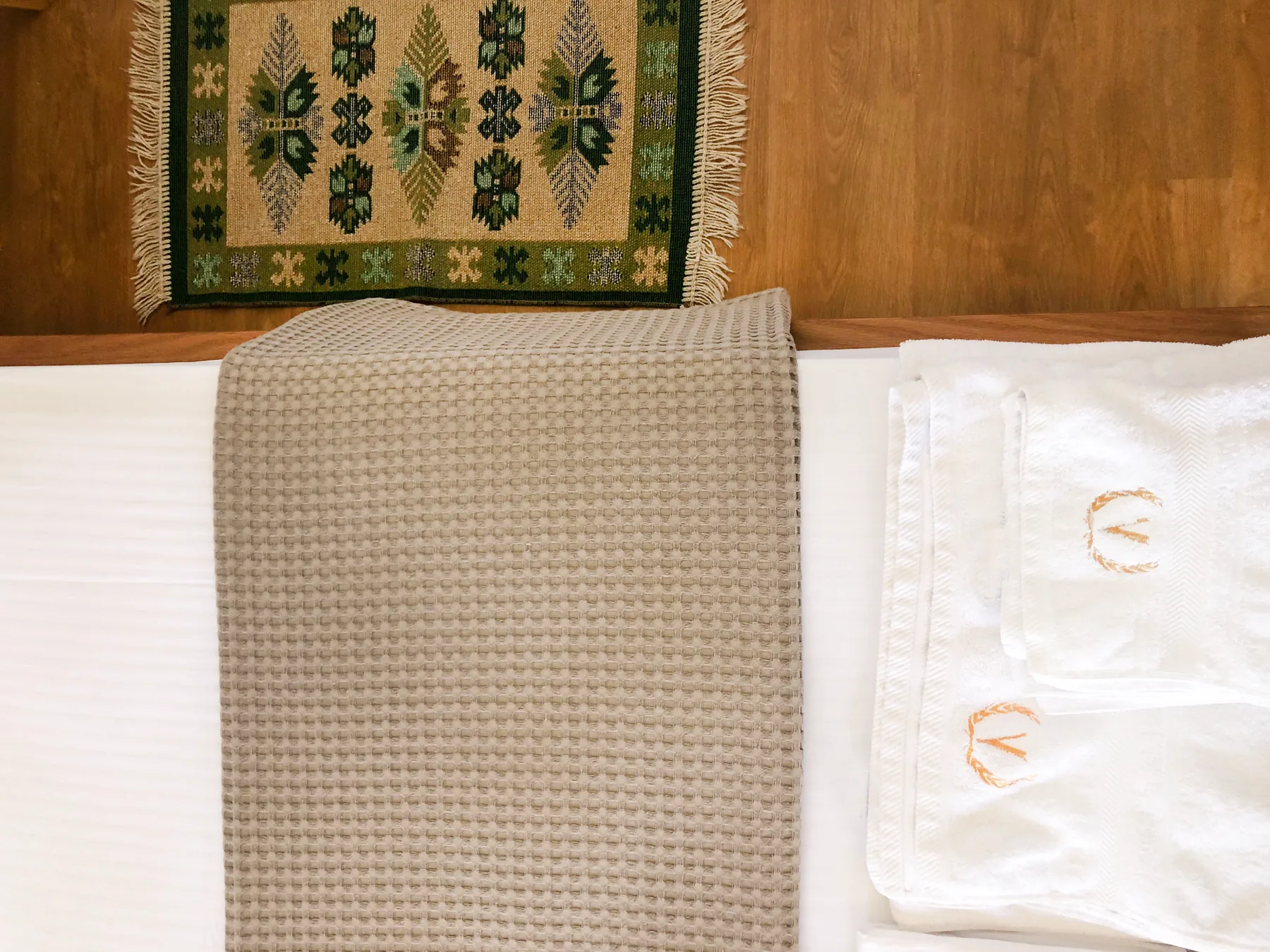 Folded towels on a white bed with a woven kilim on the timber floor beside it