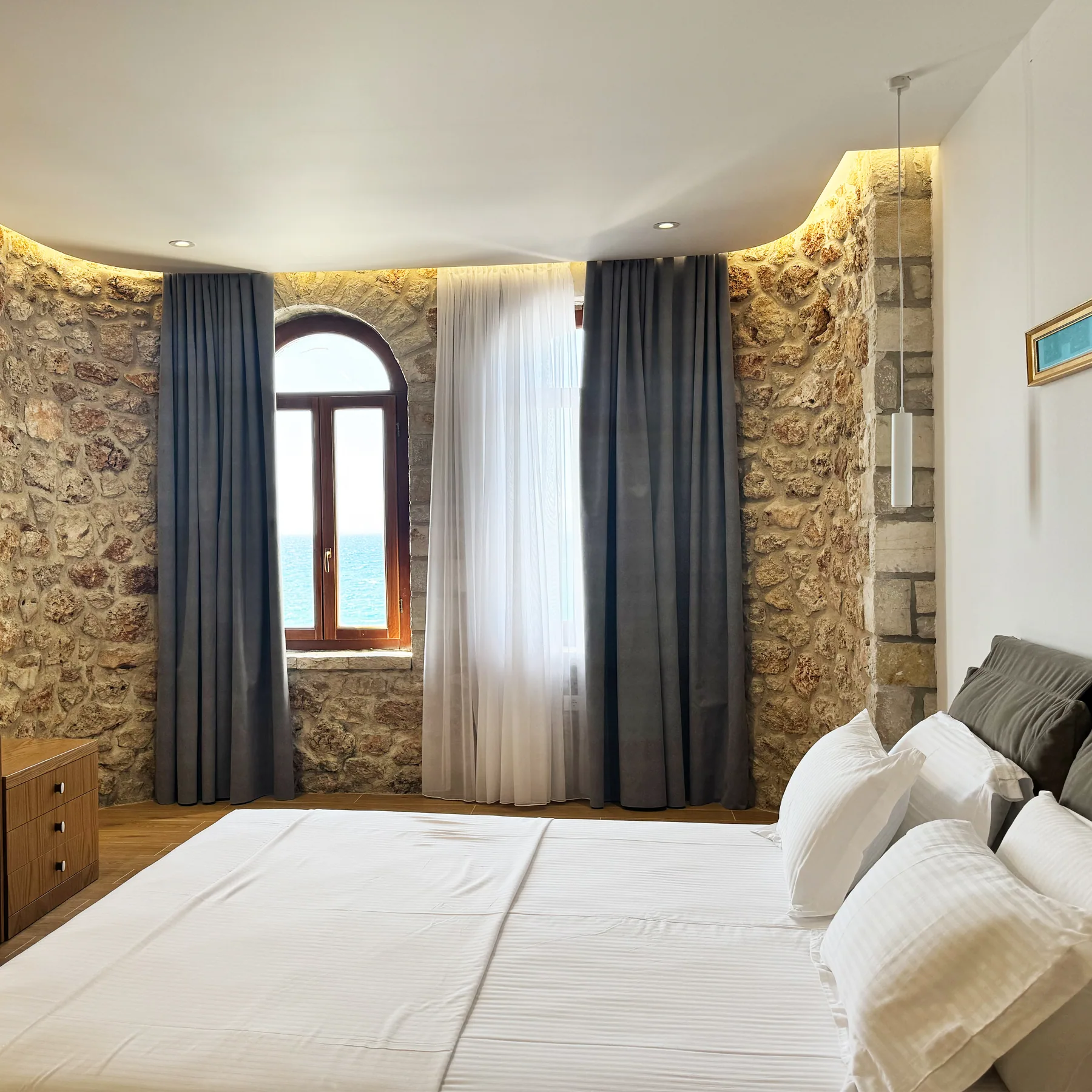 A bed beneath a small arched window in a limestone wall with a lit cornice above