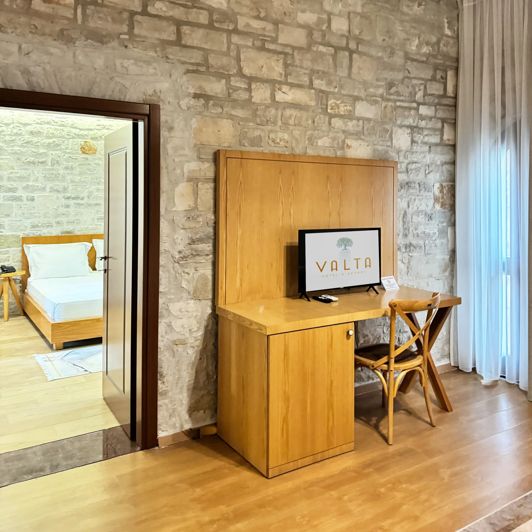 A desk and television against a limestone wall with the bedroom reflected in a mirror