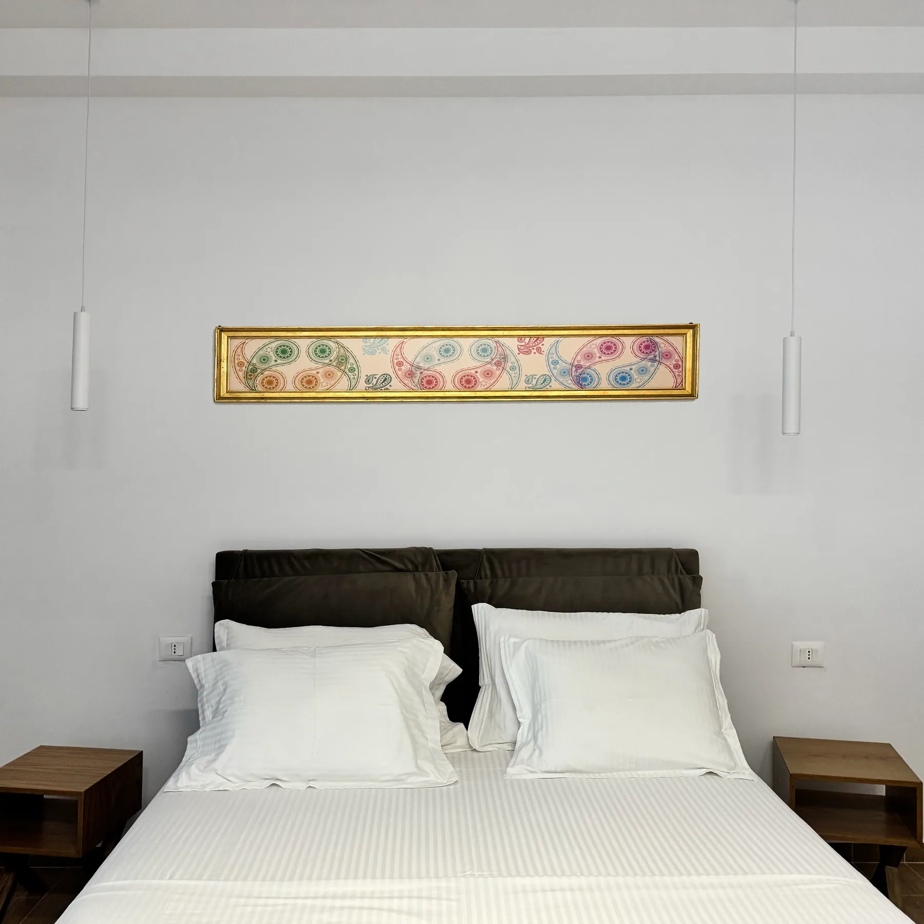 A double bed with a dark upholstered headboard beneath a long framed panel on a white wall