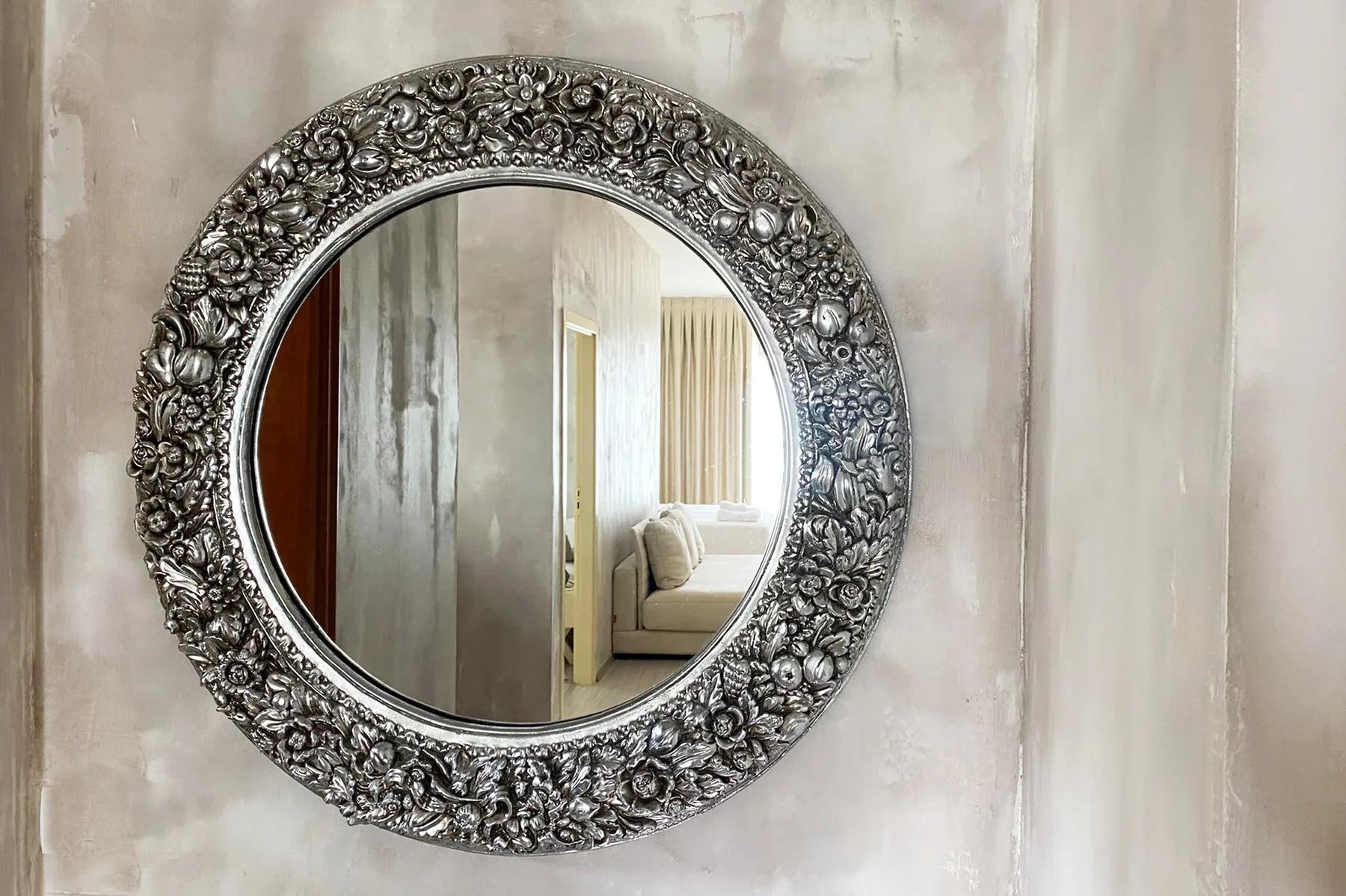 An ornate round silver mirror on a plaster wall reflecting the room behind