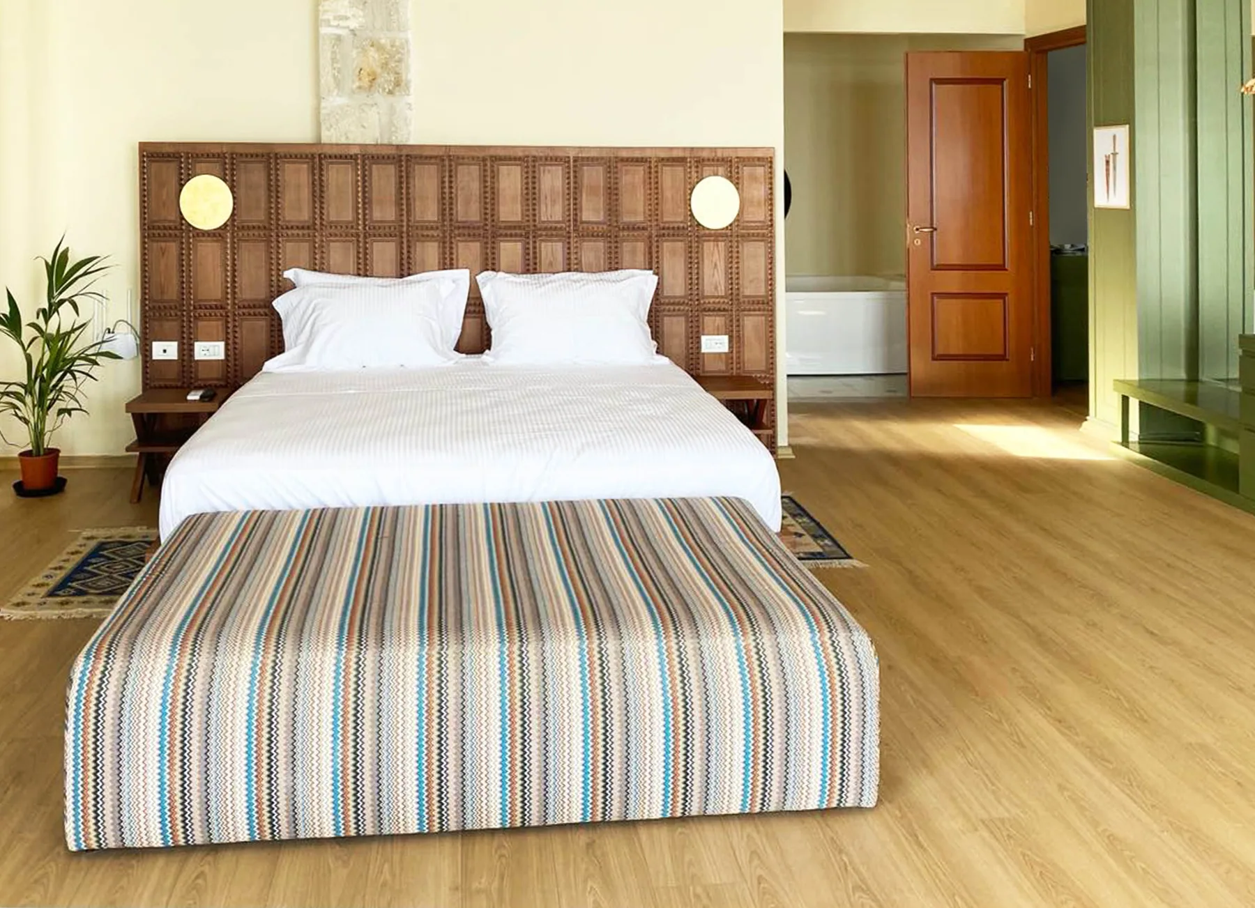 A king bed with a carved timber headboard, a striped runner and a bathroom door standing open