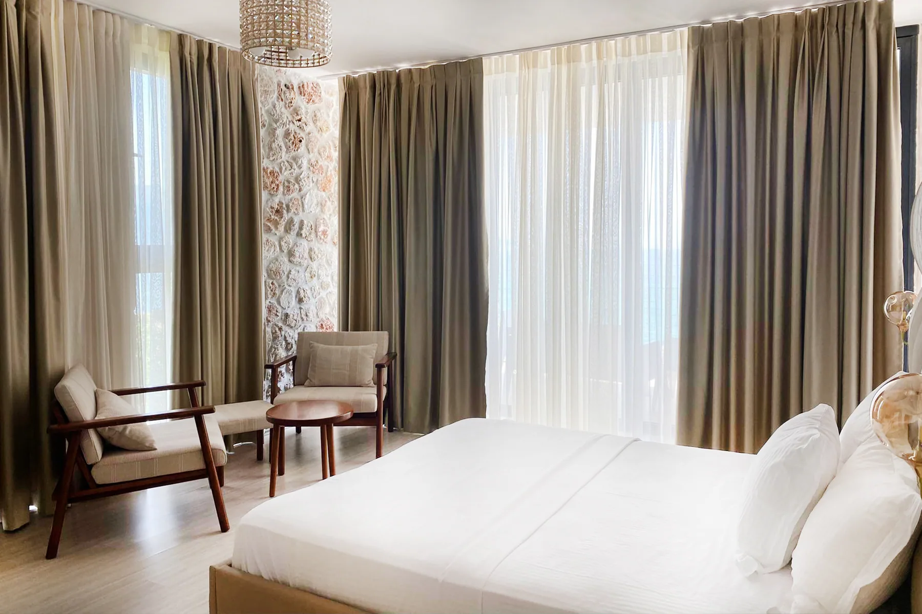 A bed and two armchairs beside full-height curtains and a limestone pier