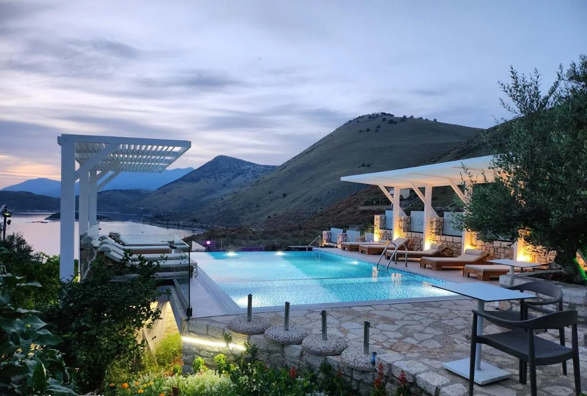 The pool at dusk with white pergolas, lit loungers and the bay pale behind the headland